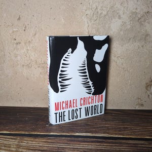 May include: A hardcover book titled The Lost World by Michael Crichton. The cover features a high-contrast black and white graphic of a dinosaur jaw with sharp teeth. The author's name is printed in red, and the title is in black text.
