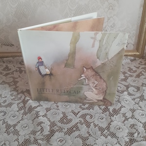 May include: A children's book titled "Little Red-Cap" with an illustration of a girl in a red hooded cloak and a wolf sitting on a rock. The book cover is a soft, light brown color with a watercolor-style illustration.