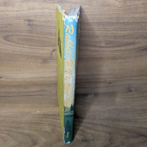 May include: The spine of a hardcover book titled Rabbit Hill by Robert Lawson. The spine features a light blue sky background transitioning to a green landscape at the bottom, with yellow text. The publisher name Viking is printed at the base.
