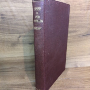 May include: A maroon-colored book titled "Elements of Radio Communication" by Morecroft. The book has gold lettering and a gold-colored top edge. The book is standing upright on a wooden surface.