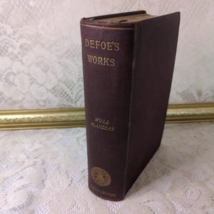 May include: A vintage hardcover book with a dark brown cover. The book's spine features gold lettering that reads "Defoe's Works" and "Moll Flanders." The book has a gold-colored top edge and a gold emblem on the bottom.