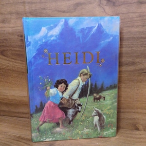 May include: A hardcover book titled HEIDI with a blue cover featuring an illustration of a child in a pink skirt and blue vest holding flowers, a child in a white shirt, and two goats in a grassy mountain meadow with blue peaks in the background.