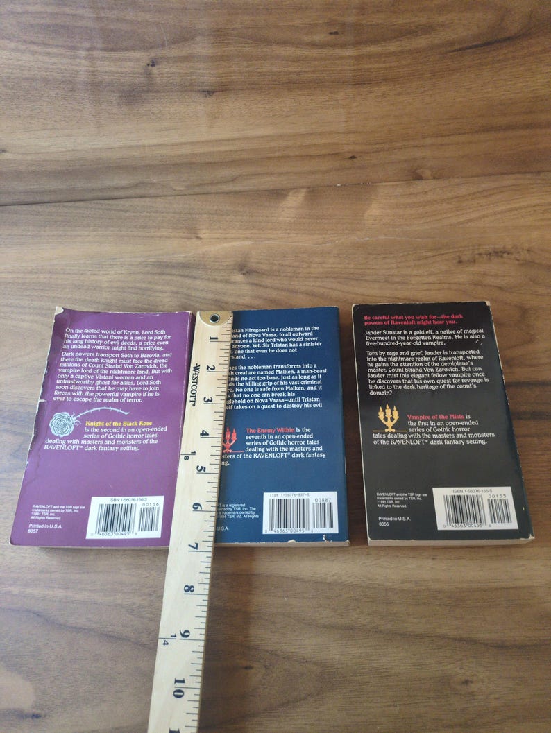 Puede incluir: Se muestran tres libros de bolsillo, con t&iacute;tulos como "Knight of the Black Rose" y "Vampire of the Mists". Una regla de madera se coloca junto a los libros. Los libros forman parte de la serie de fantas&iacute;a oscura RAVENLOFT.