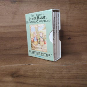 May include: A miniature book set titled "The Original Peter Rabbit Miniature Collection 5" by Beatrix Potter. The box is light green with illustrations of Peter Rabbit characters. The set includes several small books.