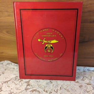 May include: A red book with a black border and gold lettering. The cover features a circular emblem with text that reads "Lu Lu Temple A.A.O.N.M.S. 100th Anniversary Plymouth Meeting, PA 1904-1904".