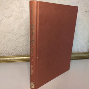 May include: A hardcover book titled "The Illustrated Brontës of Haworth" by Brian Wilks. The book has a solid reddish-brown cover with the title and author's name printed vertically on the spine in gold lettering. The book is standing upright.