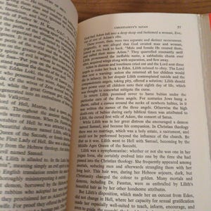 May include: An open book with text discussing Christianity's Satan and the hierarchy of Hell. The pages are aged, with black text. The book rests on a wooden surface.