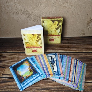 May include: A deck of Healing with the Fairies oracle cards, a matching guidebook, and a rectangular storage box. The cards feature colorful illustrations and the text Happily Ever After. The box and guidebook display fairy artwork and the title Healing with the Fairies Oracle Cards by Doreen Virtue, PhD.