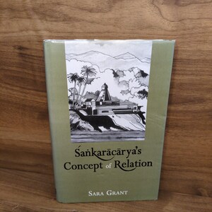 May include: A book titled "Śaṅkarācārya's Concept of Relation" by Sara Grant. The book has a light green cover with a black and white illustration of a temple and palm trees. The background is a wooden surface.