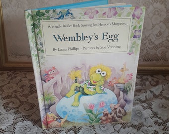 May include: A book cover featuring a green Fraggle character named Wembley from the Jim Henson's Muppets show "Fraggle Rock". The book title is "Wembley's Egg" and the author is Laura Phillips. The book cover is illustrated with a colorful cartoon style.