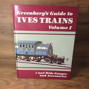 May include: A hardcover book titled "Greenberg's Guide to IVES TRAINS Volume I" with a maroon cover and a light green background. The cover features a blue and gold toy train on tracks. The text "1 and Wide Gauges and Accessories" is also visible.