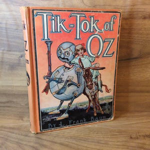 May include: Vintage hardcover book titled "Tik-Tok of Oz" by L. Frank Baum. The cover features a colorful illustration of a mechanical man, a young girl, and a donkey. The book has a faded orange cover and is set against a wooden background.