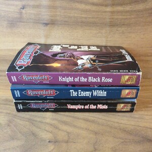 Puede incluir: Una pila de tres libros de terror de fantas&iacute;a Ravenloft. El libro superior se titula "Knight of the Black Rose" con una portada morada. El libro del medio es "The Enemy Within" con una portada azul. El libro inferior es "Vampire of the Mists".