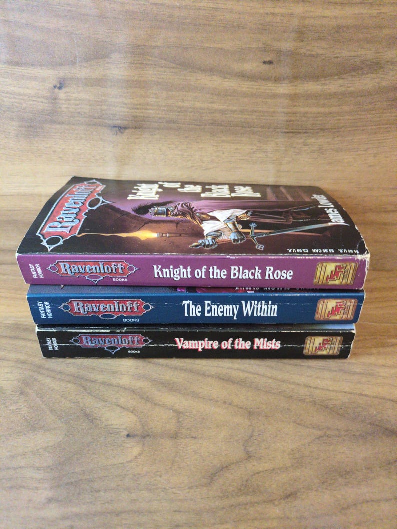 Puede incluir: Una pila de tres libros de terror de fantas&iacute;a Ravenloft. El libro superior se titula "Knight of the Black Rose" con una portada morada. El libro del medio es "The Enemy Within" con una portada azul. El libro inferior es "Vampire of the Mists".
