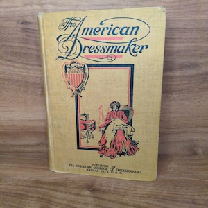 May include: A vintage book titled "The American Dressmaker" with a tan cover. The cover features ornate script and a framed illustration of a person sewing. The book is published by The American College of Dressmaking, Kansas City, U.S.A.