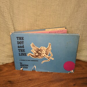 May include: A vintage book titled "THE DOT and THE LINE" by Norton Juster. The cover is light blue with an illustration of a cherub with a bow and arrow. The book is on a wooden surface.
