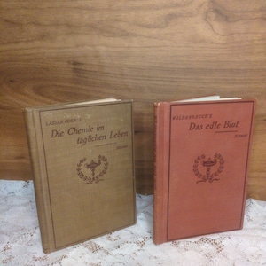 May include: Two vintage books with German titles. One book is tan with the title "Die Chemie im täglichen Leben" and the other is red with the title "Das edle Blut". Both books have a decorative emblem.