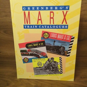 May include: A yellow book titled "GREENBERG'S MARX TRAIN CATALOGUES" with red and blue text. The book features images of vintage train catalogs from Louis Marx & Co., showcasing train illustrations and catalog covers.