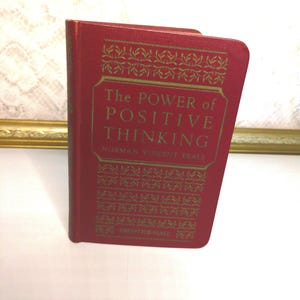 May include: A red hardcover book titled "The Power of Positive Thinking" by Norman Vincent Peale. The book's cover features gold lettering and decorative patterns. The book is slightly angled, revealing its spine and pages.