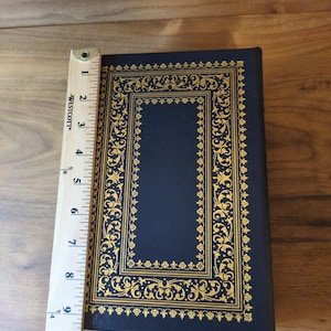 May include: A navy blue book with gold filigree detailing. The book measures approximately 9 inches in length. The cover features an ornate rectangular border and decorative corner elements.