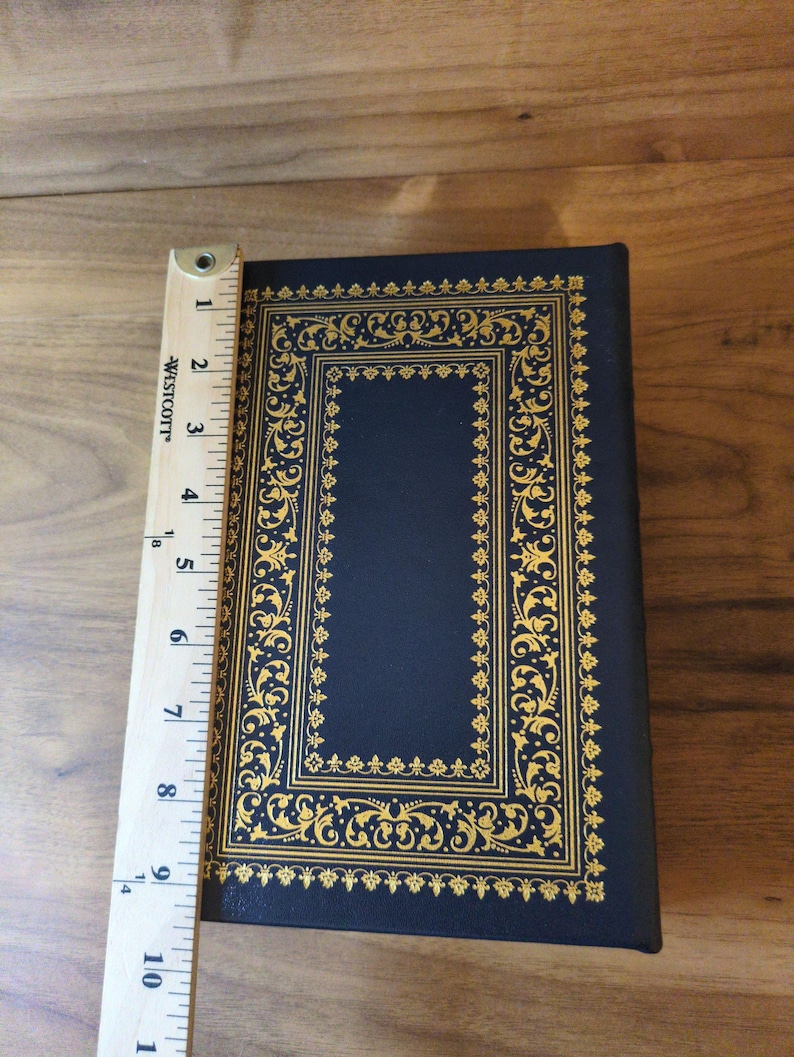 May include: A navy blue book with gold filigree detailing. The book measures approximately 9 inches in length. The cover features an ornate rectangular border and decorative corner elements.