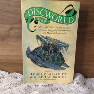 May include: A vintage copy of "The Discworld Mapp" by Terry Pratchett and Stephen Briggs. The book cover features a green banner with the title, and an illustration of a turtle carrying a flat world. The text reads "Being the Onlie True & Mostlie Accurate Mappe of the Fantastyk and Magical Dyscworlde."