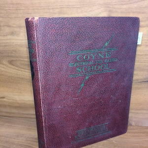 May include: A vintage book with a burgundy cover featuring the text "COYNE ELECTRICAL AND RADIO SCHOOL" in green. The cover has a textured pattern and a lightning bolt graphic. The book is slightly angled, showcasing its spine and front cover.