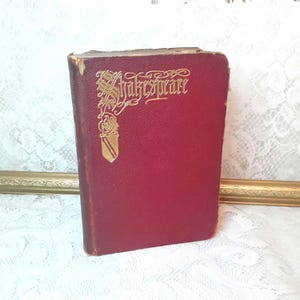 May include: A small, antique book with a deep red cover. The gold lettering on the cover reads "Shakespeare" in an ornate font. The book's edges show signs of wear, suggesting its age and use. The book is standing upright.