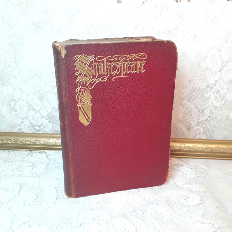 May include: A small, antique book with a deep red cover. The gold lettering on the cover reads "Shakespeare" in an ornate font. The book's edges show signs of wear, suggesting its age and use. The book is standing upright.