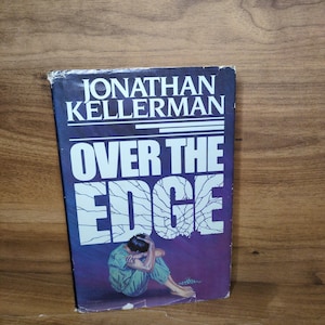 May include: A paperback book titled Over the Edge by Jonathan Kellerman. The cover features a dark blue and purple gradient background with white text and a graphic of a person sitting in a fetal position, wearing light green clothing.
