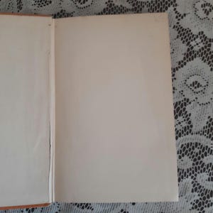 K&ouml;nnte beinhalten: Aufgeschlagenes Buch mit gealterten, cremefarbenen Seiten. Der Einband ist auf der linken Seite sichtbar, mit einem Hauch von Orange. Die Seiten sind leer, was auf ein altes Buch hindeutet. Der Hintergrund ist eine wei&szlig;e Spitzendecke.