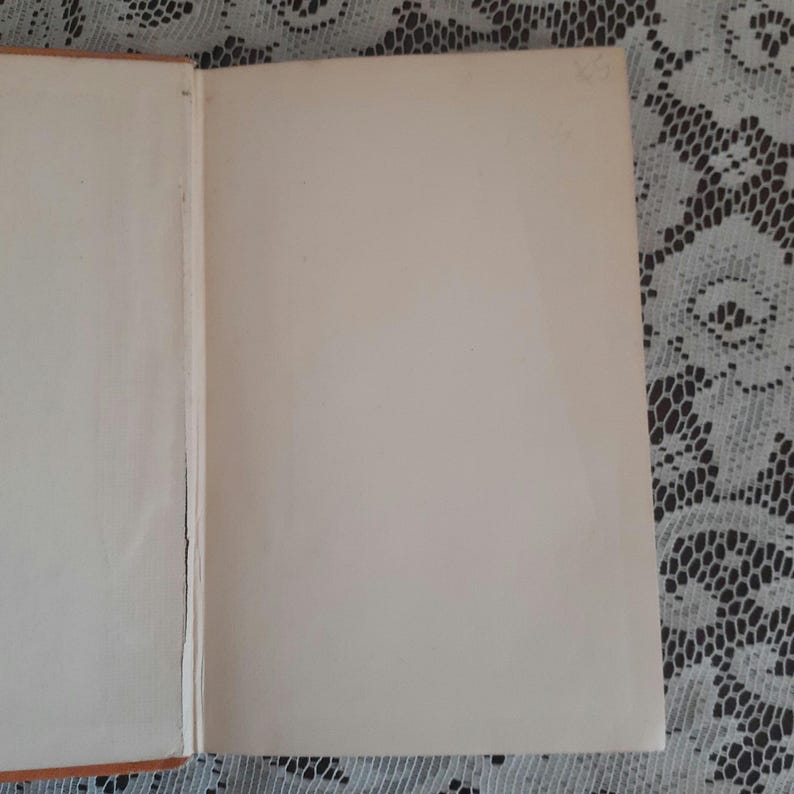 K&ouml;nnte beinhalten: Aufgeschlagenes Buch mit gealterten, cremefarbenen Seiten. Der Einband ist auf der linken Seite sichtbar, mit einem Hauch von Orange. Die Seiten sind leer, was auf ein altes Buch hindeutet. Der Hintergrund ist eine wei&szlig;e Spitzendecke.