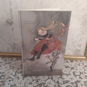 May include: The Legend of Rosepetal book cover features a watercolor illustration of a woman with long blonde hair, wearing a red dress and black shoes, floating above a rose bush. The book title is written in a stylized font.