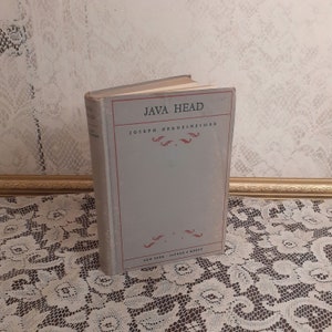 May include: A vintage book titled "JAVA HEAD" by Joseph Hergesheimer. The book has a light gray cover with red accents and text. Published by Alfred A. Knopf in New York. The book is displayed on a white lace tablecloth.