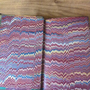 May include: An open book featuring marbled endpapers with a repeating wavy pattern in shades of deep red, blue, yellow, and white. The paper displays a complex, swirling design characteristic of traditional marbling techniques on the interior covers.