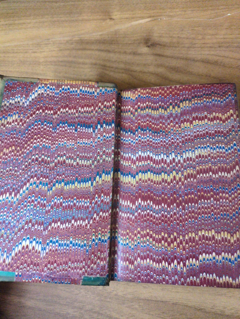 May include: An open book featuring marbled endpapers with a repeating wavy pattern in shades of deep red, blue, yellow, and white. The paper displays a complex, swirling design characteristic of traditional marbling techniques on the interior covers.