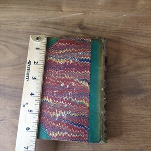 May include: A vintage hardcover book with a marbled paper cover in red, blue, and yellow, featuring a green leather spine and corner accents. A Westcott ruler is placed alongside the book, showing a height of approximately 7.5 inches.