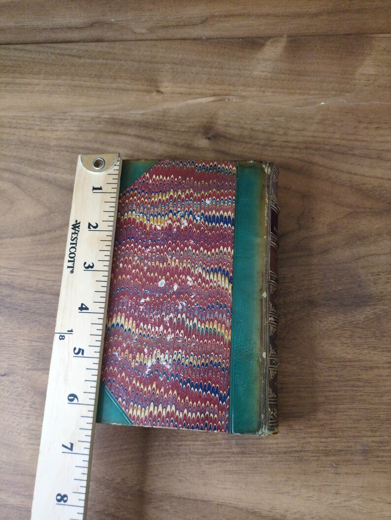 May include: A vintage hardcover book with a marbled paper cover in red, blue, and yellow, featuring a green leather spine and corner accents. A Westcott ruler is placed alongside the book, showing a height of approximately 7.5 inches.