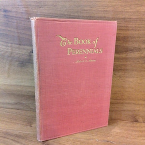 May include: A vintage book titled "The Book of Perennials" with a coral-colored cover and gold lettering. The book is standing upright on a wooden surface, showcasing its title and author, Alfred C. Hottes.