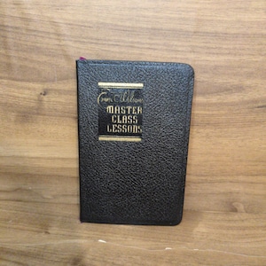May include: A small, black book with the title "Master Class Lessons" in gold lettering. The book has a textured cover and a purple ribbon bookmark. The book is on a wooden surface.