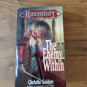 Puede incluir: Un libro de bolsillo titulado "The Enemy Within" de Christie Golden. La portada presenta a un hombre con un abrigo rojo con botones dorados y una espada, y el reflejo de un rostro amenazante. El libro es parte de la serie Ravenloft.