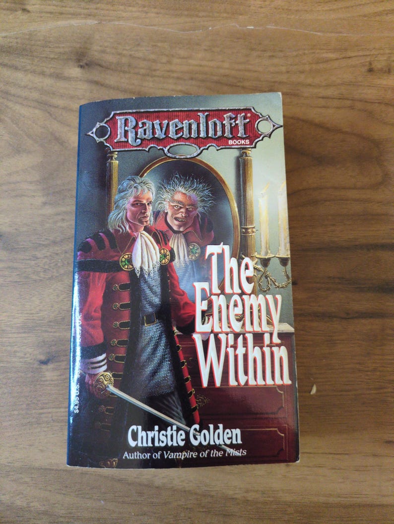 Puede incluir: Un libro de bolsillo titulado "The Enemy Within" de Christie Golden. La portada presenta a un hombre con un abrigo rojo con botones dorados y una espada, y el reflejo de un rostro amenazante. El libro es parte de la serie Ravenloft.