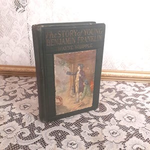 May include: A green hardcover book titled "The Story of Young Benjamin Franklin" by Wayne Whipple. The cover features an illustration of a man in a colonial-era outfit pointing at a tree with a young boy standing nearby.