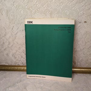 May include: An IBM System/360 COBOL programming manual. The book has a green cover with white text and a white border. The text reads "SYSTEM/360 COBOL Writing Programs in COBOL Test" and "Programmed Instruction Course."