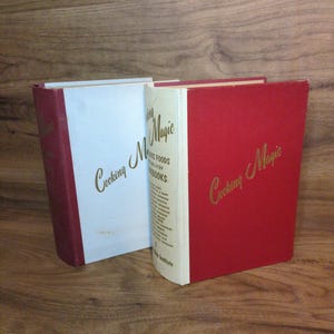 May include: Two vintage cookbooks titled "Cooking Magic" are displayed. One book has a white cover with a red spine, the other is red with a gold title. The books are standing upright on a wooden surface.