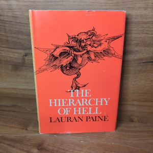 May include: A vintage paperback book titled "The Hierarchy of Hell" by Lauran Paine. The book has a bright orange cover with a black illustration of a demonic figure. The book is standing upright on a wooden surface.