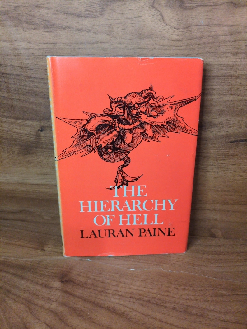 May include: A vintage paperback book titled "The Hierarchy of Hell" by Lauran Paine. The book has a bright orange cover with a black illustration of a demonic figure. The book is standing upright on a wooden surface.