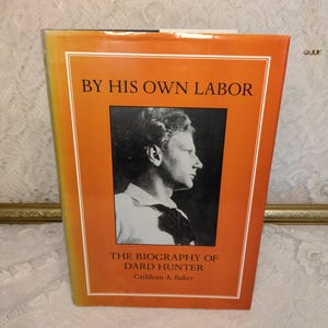 May include: A hardcover book titled "BY HIS OWN LABOR" with an orange cover and white border. The book features a black and white portrait of a man and the text "THE BIOGRAPHY OF DARD HUNTER" and "Cathleen A. Baker".