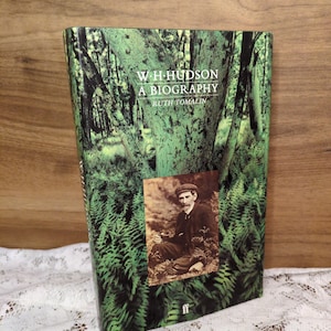 May include: A hardcover book titled "W.H. Hudson A Biography" by Ruth Tomalin. The cover features a green forest scene with ferns and trees. A sepia-toned photograph of a man in a cap is on the front. The book is displayed on a white lace surface.