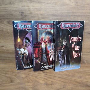 Puede incluir: Se muestran tres libros de Ravenloft, con ilustraciones g&oacute;ticas y t&iacute;tulos como "Vampire of the Mists". Las portadas presentan im&aacute;genes oscuras con figuras y texto, sobre un fondo de madera. Los autores son James Lowder y Christie Golden.